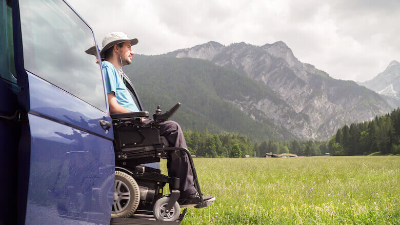 Campingfahrzeug mit Rollstuhllift parkt auf einer Wiese mit einer Berglandschaft im Hintergrund.